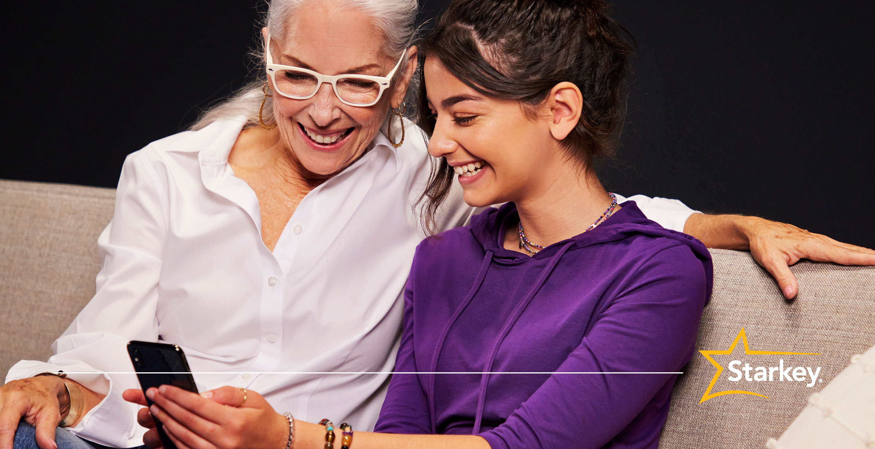 Senior woman sitting on a couch with arm around younger woman looking at smartphone